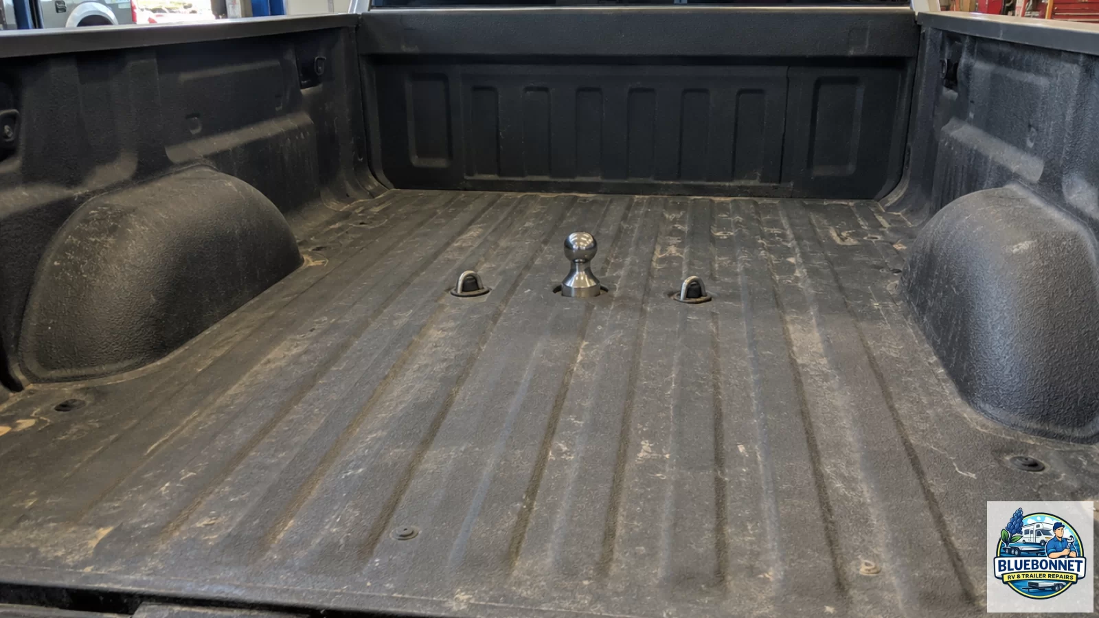 Tailgate view into a pickup bed showing an under-bed gooseneck ball with safety-chain U-bolt loops located between the wheel wells in the proper towing position